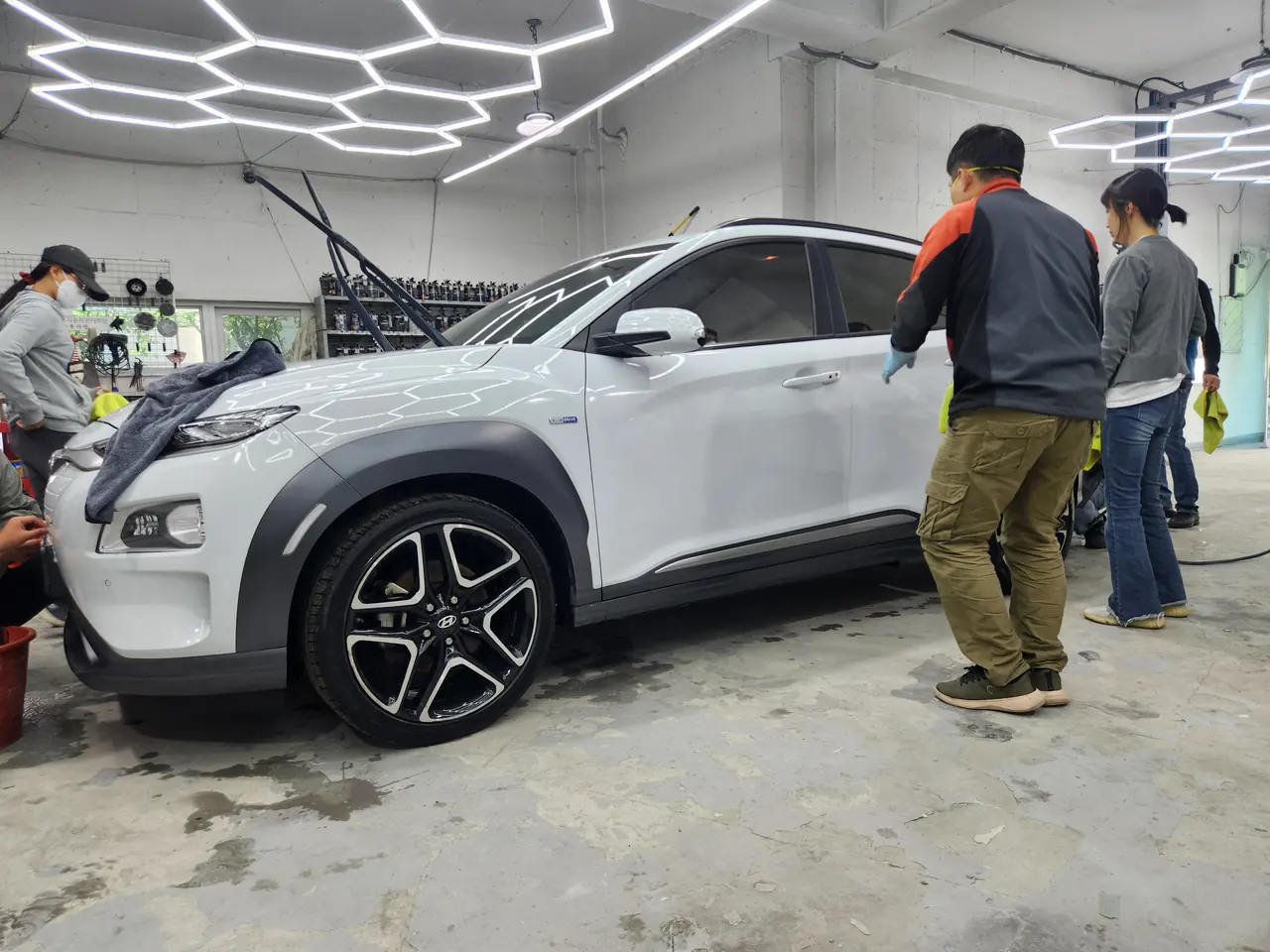 흰색 SUV 측면 앞에서 강사가 교육생 옆에 서서 도장면을 함께 살피는 장면, 육각 조명이 면에 그대로 비친다